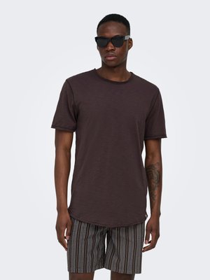 Uomo con occhiali da sole neri, camicia a maniche corte marrone scuro e pantaloncini a righe bianche e nere, in piedi davanti a uno sfondo semplice.