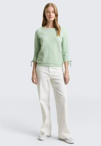Lichtgroene textuursweatshirt met driekwartmouwen en strikaccenten. Gecombineerd met witte wijde broekspijpen en casual schoenen.