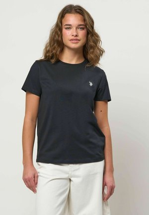 Mladá žena s vlnitými vlasmi, oblečená v čiernom tričku s krátkym rukávom s malým bielym logom a bielych nohaviciach, stojaca pred jednoliatym pozadím.