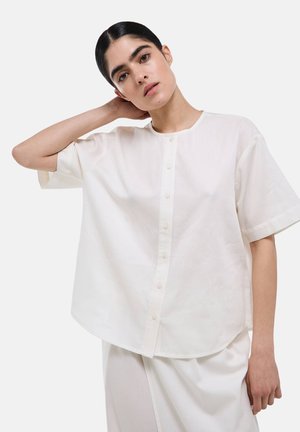 Femme aux cheveux foncés portant une chemise blanche ample à manches courtes boutonnée et une jupe blanche, debout devant un fond uni.