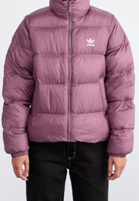 Lila puffjacka med hög krage, dragkedja fram och vit adidas-logga på vänster bröst. Har kviltat mönster och elastiska ärmslut.