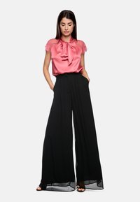 Rosa Satinbluse mit Spitzeärmeln und Schleifenkragen, kombiniert mit schwarzen weit geschnittenen Hosen mit Taschen und einem fließenden Design.