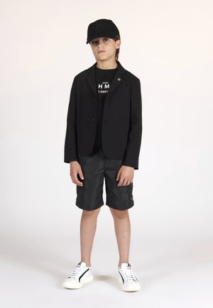 Blazer nero su una t-shirt nera, pantaloni corti neri e sneakers bianche. Il cappellino è anch'esso nero con un logo in rilievo.
