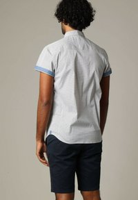 Korte mouwen shirt in het wit met een geometrisch patroon, blauwe accenten bij de kraag en mouwsluiten. Gecombineerd met donkerblauwe shorts.