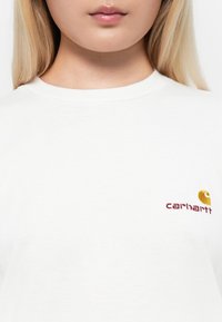 Hvid bomulds t-shirt med rund hals, med et gult og burgunder broderet Carhartt logo på venstre brystområde.