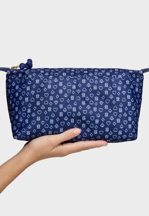 Mano sosteniendo un pequeño bolso con cremallera azul marino con un patrón de palos de cartas blancos y un detalle de tirador de cremallera con cuentas.