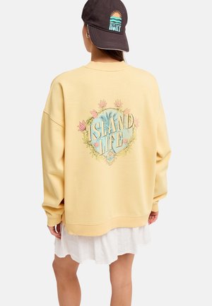 Personne portant un sweat-shirt jaune avec un motif floral "Island Life" dans le dos, une jupe blanche et une casquette noire avec un logo soleil et mer.