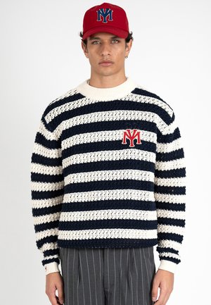 Hombre con gorra roja y jersey de punto a rayas azul marino y blanco con logo rojo "NY" y pantalones gris con rayas finas sobre fondo blanco.