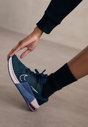 Chaussure de sport avec une tige en maille texturée bleu foncé, logo swoosh blanc, et une semelle bleu clair avec des accents violets. Chaussette noire visible.