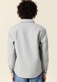 Veste chemise en mélange de laine grise, avec un col, des poignets à bouton et un ourlet arrondi. Texture douce et détails de couture soignés.