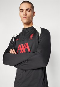 Czarny sportowy top z wysokim kołnierzem, ozdobiony czerwonym logo Liverpool FC oraz brandingiem "AXA". Posiada białe akcenty na ramionach i gładką teksturę.