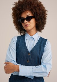 Donna con i capelli ricci che indossa occhiali da sole neri, orecchini a cerchio argento, una camicia abbottonata azzurro chiaro e un gilet senza maniche blu scuro, con le braccia incrociate.