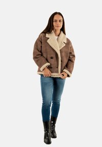Chaqueta de borreguillo marrón con diseño de doble botonadura, cuello y puños en color crema, combinada con vaqueros azules y botas negras.