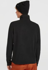 Sort fleece-pullover med høj krave, lange ærmer og en glat tekstur. Båret sammen med en sort hue, der viser minimal branding på hatten.