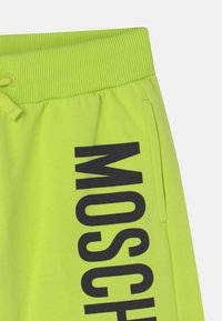 Neonově zelené tepláky s ribbeným pasem, detailem na šňůrku, boční kapsou a velkým černým nápisem "MOSCH" na levé noze.