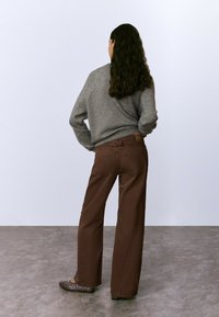 Mujer con cabello rizado y largo de pie de espaldas, usando un suéter gris, pantalones anchos marrones y planos oscuros con detalles de ojales.