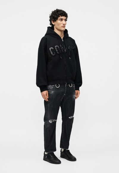 Νέος άνδρας με σγουρά μαλλιά που φοράει μαύρο φούτερ με φερμουάρ και ανάγλυφο λογότυπο, σκισμένο σκούρο τζιν με μεταλλικούς κρίκους και μαύρα αθλητικά παπούτσια.