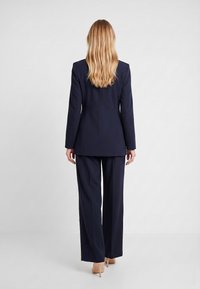 Femme aux longs cheveux blonds de dos, portant un tailleur-pantalon bleu marine et des talons beiges sur un fond uni clair.