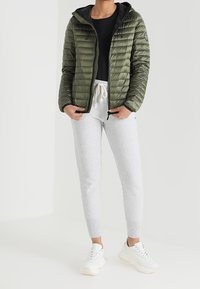 Gevoerde groene jas met een zwarte binnenkant, ritsluiting en capuchon, gecombineerd met lichtgrijze trainingsbroek en witte sneakers.