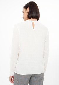 Blusa blanca de manga larga con puntos texturizados, detalle de ojal en la parte posterior y puños abotonados, combinada con pantalones grises a cuadros.