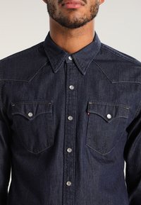 Tmavě modrá denimová košile s ostrým límcem, tiskovými knoflíky, dvěma náprsními kapsami a kontrastním prošitím. Vyznačuje se texturovanou tkaninou a západním stylem designu.
