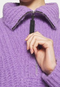 Pull-over en maille violet avec col haut et fermeture à demi-glissière. Présente un tissu texturé et un tirette de fermeture éclair noire contrastante.