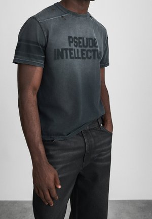 Man draagt een vervaagd zwart T-shirt met versleten gaten en de tekst "PSEUDO INTELLECTUAL", gecombineerd met een zwarte spijkerbroek, staand tegen een eenvoudige achtergrond.