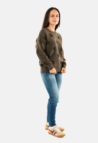 Donna in piedi che indossa un maglione verde oliva a stelle, jeans blu, calzini bianchi e sneakers beige con accenti in bordeaux e giallo.
