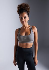 Grå ribbet sports-bh med scoop-halsutskjæring, som har tre knappdetaljer foran og en matchende elastisk bånd, kombinert med svarte tights.