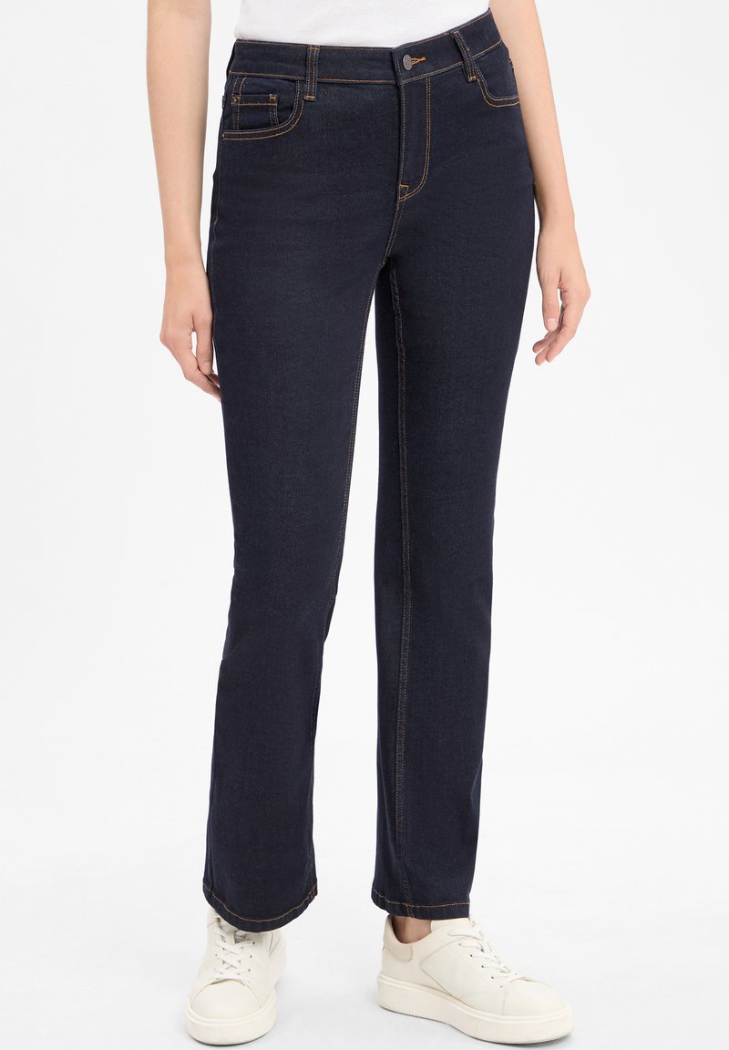 Jean en denim bleu foncé avec une coupe droite, présentant des coutures contrastées et des poches avant, accompagnés de baskets de couleur claire.