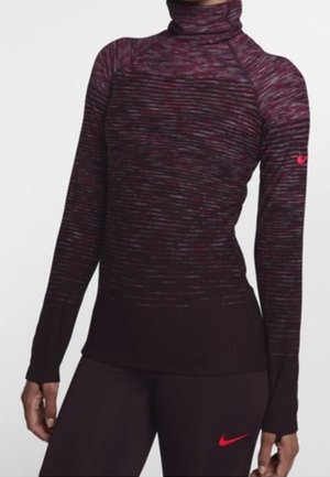 Sportliche Person trägt einen dunkelroten und schwarz gestreiften Rollkragenpullover mit langen Ärmeln und passende Leggings mit roten Nike-Logos auf Ärmel und Oberschenkel.