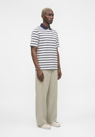 Άνδρας με κοντά ξανθά μαλλιά που φοράει λευκό και ναυτικό ριγέ πόλο πουκάμισο, μπεζ παντελόνια με φαρδιά μπατζάκια και λευκά αθλητικά παπούτσια, στέκεται μπροστά σε ένα μονόχρωμο φόντο.