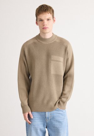Beige knit sweater with a crew neck, textured pattern, and front pocket. Features ribbed cuffs and hem, worn with light blue jeans.