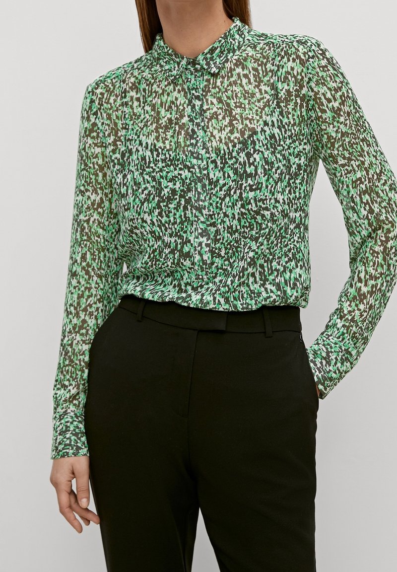 Blusa verde trasparente con un sottile motivo astratto in diverse sfumature. Presenta un colletto alto e una chiusura con bottoni davanti.