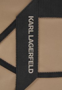 Tessuto beige con finitura testurizzata, caratterizzato da un'etichetta nera prominente impressa con "KARL LAGERFELD" in argento. Cinghie nere con dettaglio di cucitura.