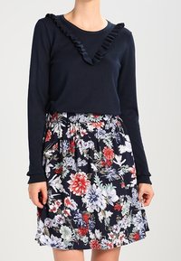 Marinblå långärmad topp med volangkrage, ihop med en blommig kjol med röda, blå och gröna blommor på en mörk bakgrund.