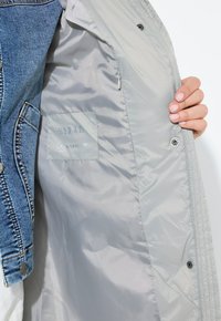 Jeansjacke mit grauem, wasserabweisendem Futter, versehen mit einem Logo-Patch, Druckknöpfen und sichtbaren Nähten entlang der Nähte.