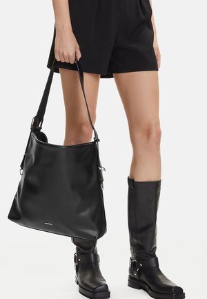 Persona vistiendo pantalones cortos negros y botas de cuero negras hasta la rodilla, sosteniendo un gran bolso de cuero negro por la correa del hombro.