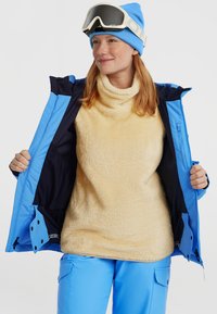 Skijack in blauw met een binnen fleece trui in beige. De fleece heeft een hoge kraag en de jack heeft contrasterende binnenpanelen.