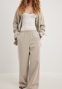 Bež široke hlače z elastiko v pasu, v kombinaciji z belim oprijetim majico brez rokavov in usklajenim bež cardiganom s trakastimi detajli.