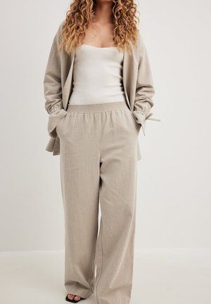 Pantaloni beige a gamba larga con cintura elastica, abbinati a una canotta bianca aderente e a una cardigan beige coordinato con dettagli a fiocco.