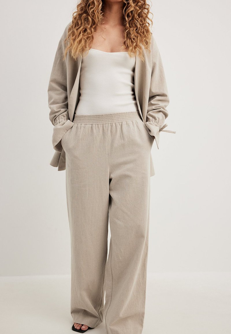 Bež široke hlače z elastiko v pasu, v kombinaciji z belim oprijetim majico brez rokavov in usklajenim bež cardiganom s trakastimi detajli.