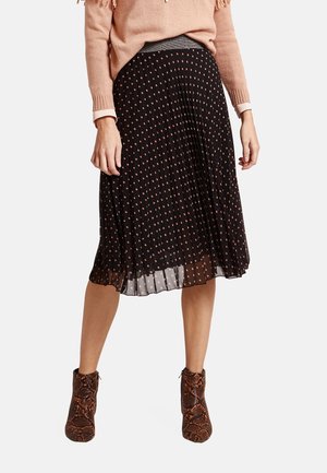 Vrouw met een zwarte plissé midi-rok met roze stippen, een lichtroze trui en bruine enkellaarsjes met slangenhuidpatroon.