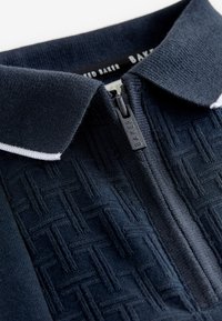 Close-up van een donkerblauw, gestructureerd stoffen poloshirt met een ritssluiting bij de kraag en witte bies, rits-trekker gegraveerd met de merknaam "BAKER".