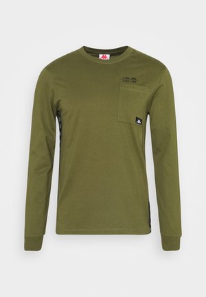 Chemise manches longues vert olive avec poche poitrine et petit patch logo noir, avec un texte subtil au-dessus de la poche.