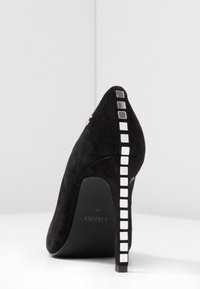 Zapato de tacón alto de ante negro con punta afilada y un acento blanco de patrón cuadrado a lo largo del tacón. Nombre de la marca "LIUJO" grabado en la suela.