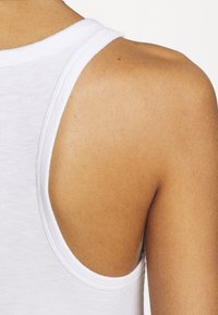 Vit linne med breda axelremmar, ribbad struktur och rundad halsringning, uppvisad mot en ljus bakgrund som avslöjar hudton.