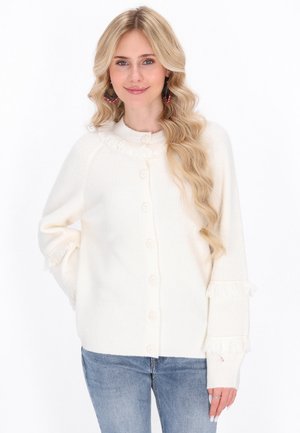 Donna bionda che indossa un maglione bianco con bottoni e dettagli in frangia, jeans blu, sorridente con una mano in tasca.