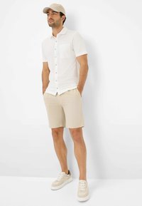 Witte kortarmige shirt met een borstzak, gecombineerd met beige shorts. Een lichtbeige pet en lichtgekleurde sneakers maken de outfit af.