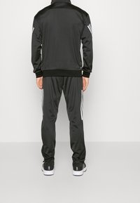Sort atletisk træningsdragt med en lynlåsjakke og joggebukser. Stoffet ser glat ud med hvide accenterstriber på siderne.